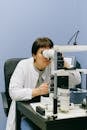 Unrecognizable female biologist in white coat watching object through microscope while sitting on chair at black table in light laboratory