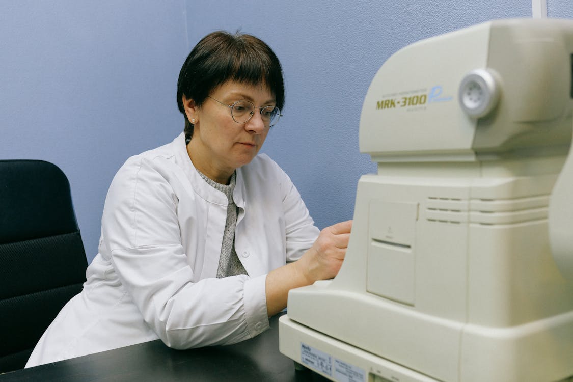 Serious middle aged eye doctor in eyeglasses working with automatic refkeratometer while sitting on chair leaned forward at black desk at work