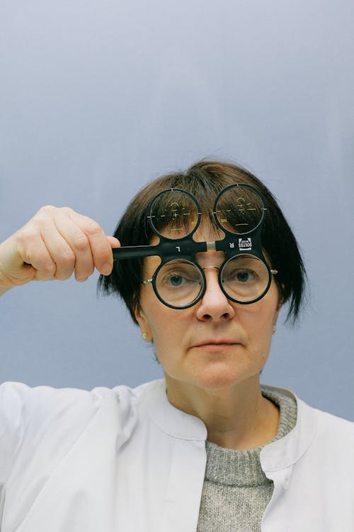 Free Female ophthalmologist in white coat watching through magnifier device for vision measurement while standing in blue background and looking at camera Stock Photo