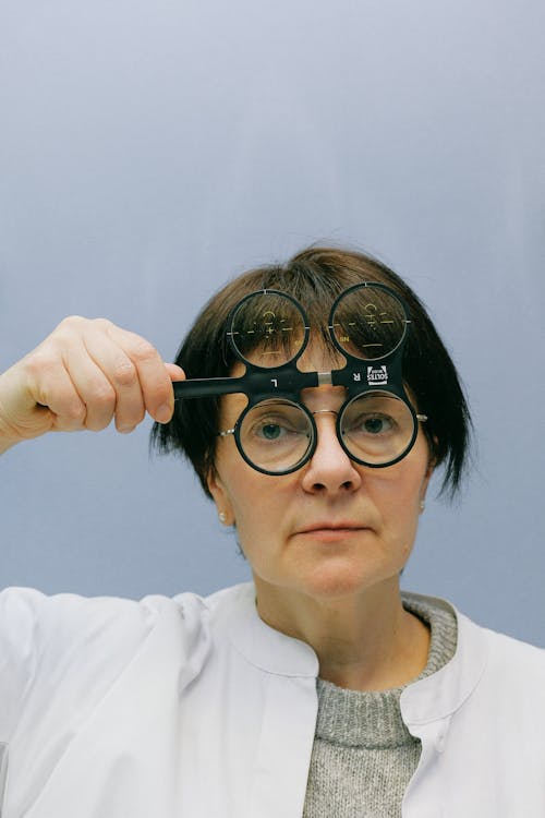 Female ophthalmologist in white coat watching through magnifier device for vision measurement while standing in blue background and looking at camera