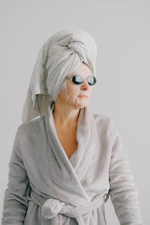 Free Aged female in gray bathrobe and with towel on head standing on gray background and looking away in sunglasses with sheet mask on face Stock Photo