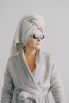 Aged female in gray bathrobe and with towel on head standing on gray background and looking away in sunglasses with sheet mask on face