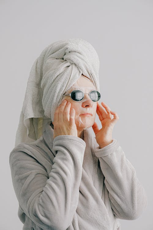 Calm woman with sheet mask on face touching face in bathrobe and towel on head while resting after bathing and looking away