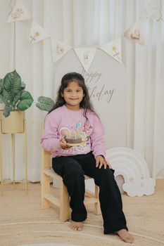 Young girl celebrating her birthday indoors with pastel decor and a small cake.