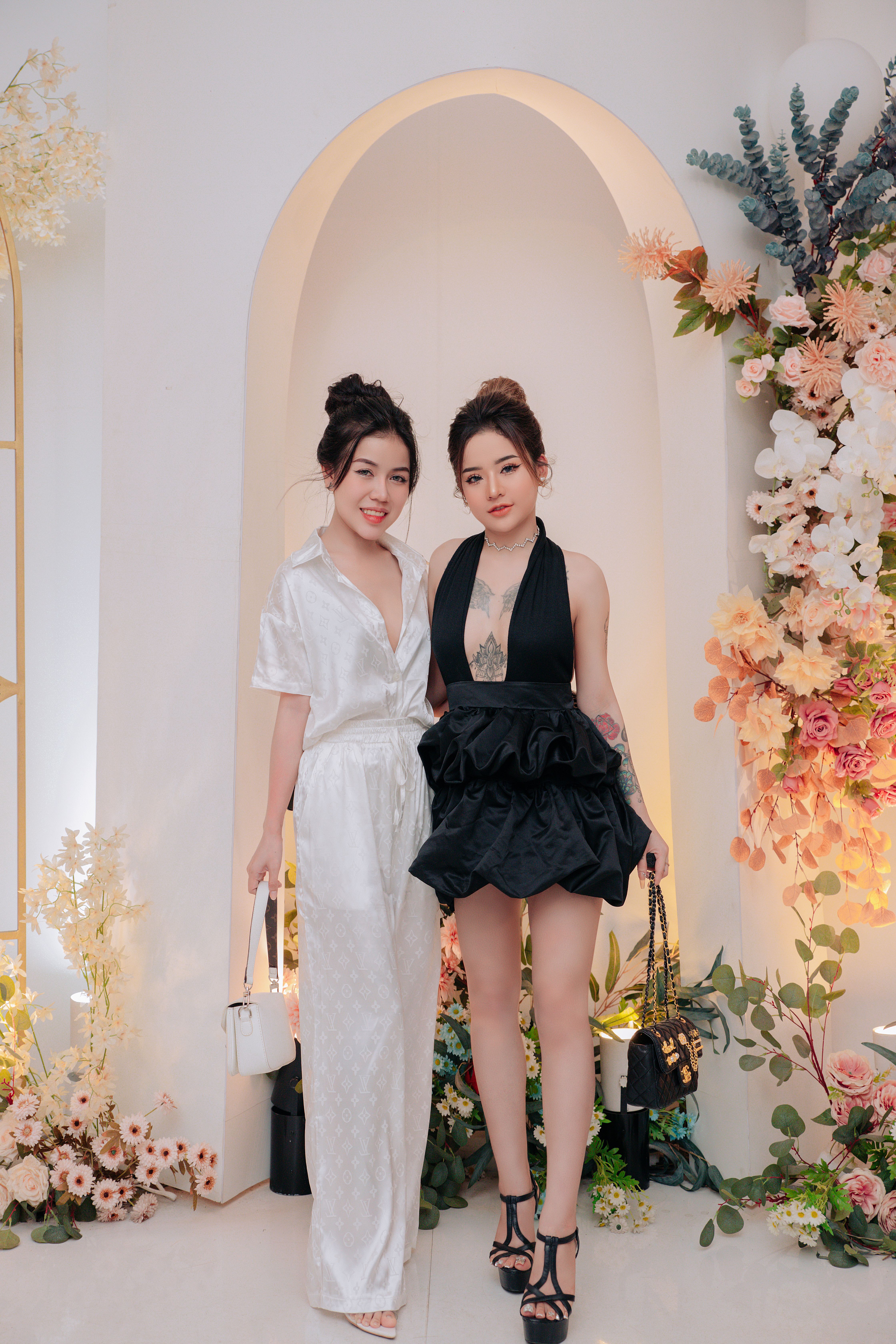 Two women in stylish outfits pose amidst floral decor in an elegant setting.