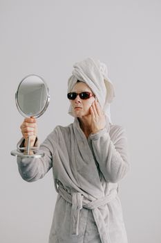 A senior woman in a bathrobe applies facial care while looking in a mirror.
