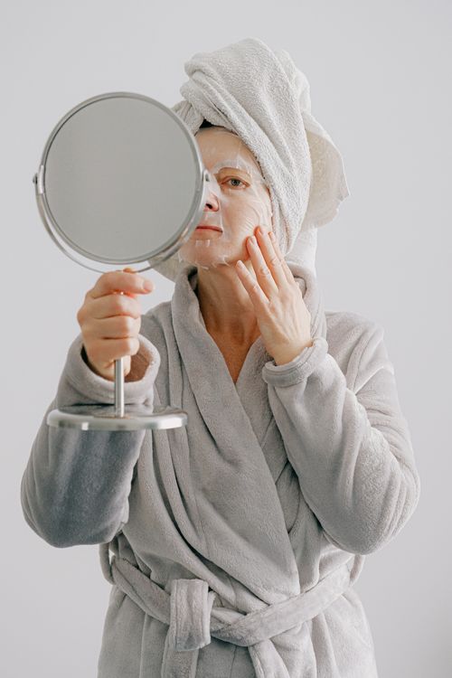 Free Woman in bathrobe applying a skincare facial mask using a mirror at home. Stock Photo