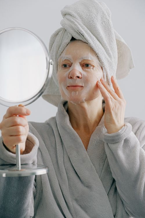 Free Adult woman in a bathrobe applying a face mask while holding a mirror indoors for skincare routine. Stock Photo