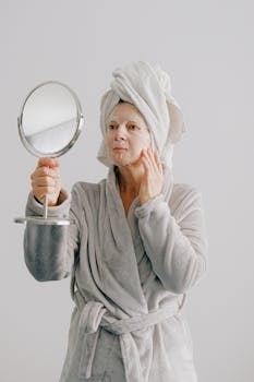 Senior woman in a bathrobe and face mask, checking her skincare in a hand mirror.