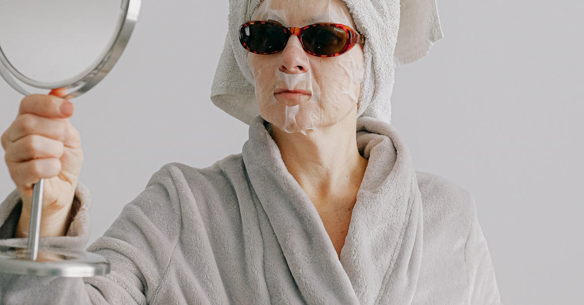 Senior female in home wear with towel on head standing against gray background after shower at home in sheet mask and sunglasses while holding table mirror in hands and looking in mirror