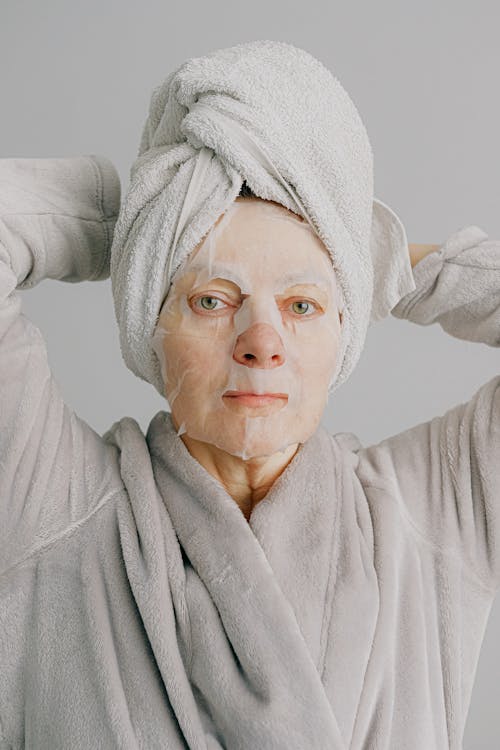 Aged relaxed female in warm bathrobe and towel on hand looking at camera and enjoying fresh sheet mask on face while standing with hands behind head