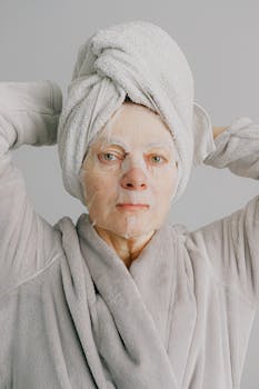 Aged relaxed female in warm bathrobe and towel on hand looking at camera and enjoying fresh sheet mask on face while standing with hands behind head