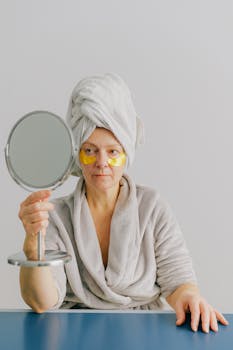 Pensive female in bathrobe with white towel on head sitting at table after bath at home with gold eye patches under eyes while holding table mirror in hands and looking in mirror