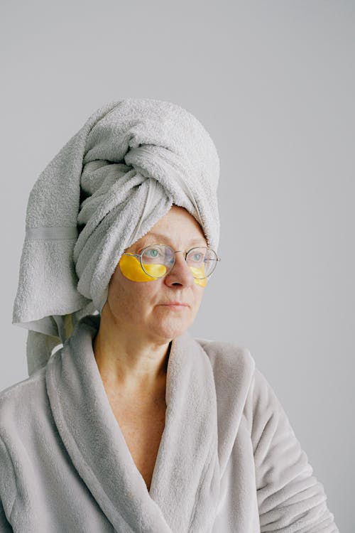 Free Pensive female in bathrobe with towel on head standing on gray background in eye patches under eyes and eyeglasses while looking away Stock Photo