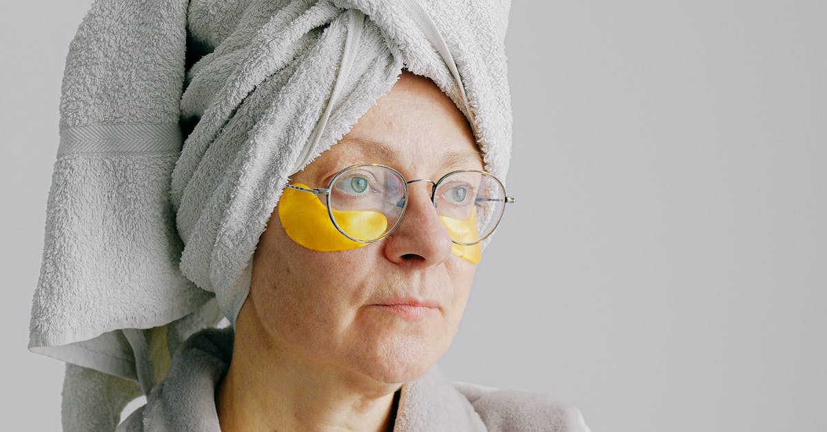 Pensive female in bathrobe with towel on head standing on gray background in eye patches under eyes and eyeglasses while looking away