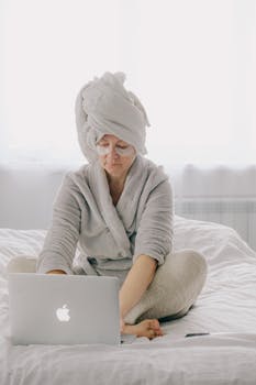 Focused female in home clothes towel on head and eye patches sitting on bed and browsing laptop while resting during weekend at home