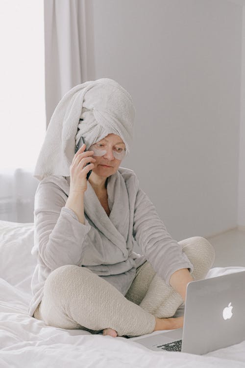 Concentrated woman in home wear sitting on bed with legs crossed and using laptop while talking on phone with towel on head and white eye patches under eyes in cozy bedroom at home