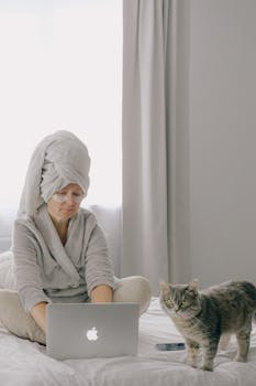 一位女士身着长袍，裹着毛巾，在床上用笔记本电脑工作，旁边有只猫，营造出舒适的家庭办公氛围