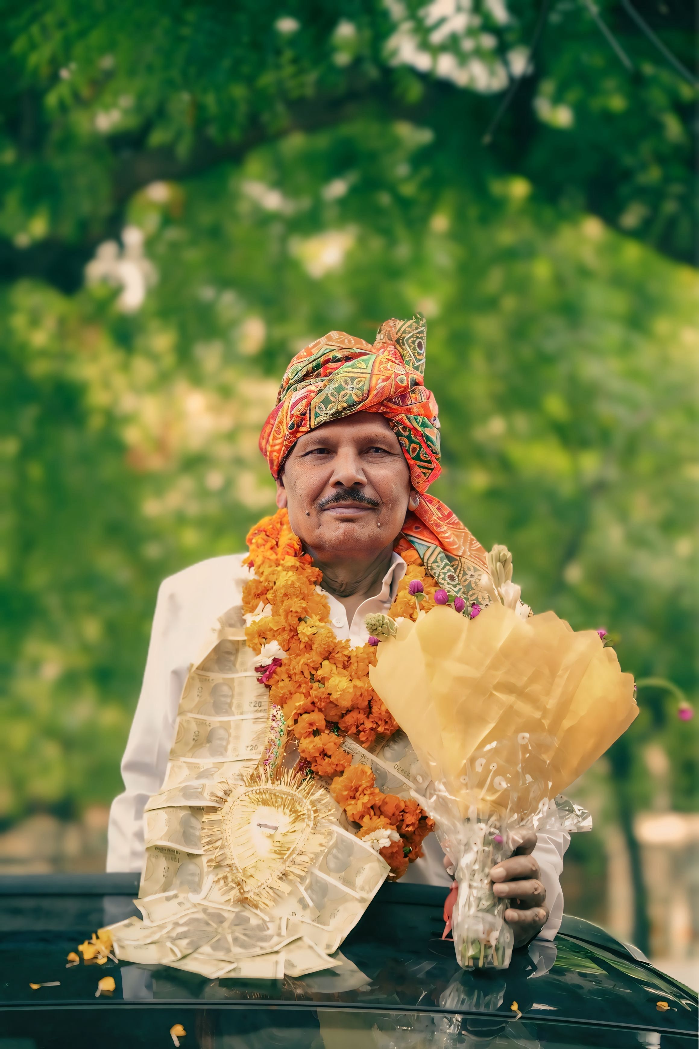 Smiling Indian man adorned with flowers and ornate turban, holding a bouquet against a lush outdoor backdrop.