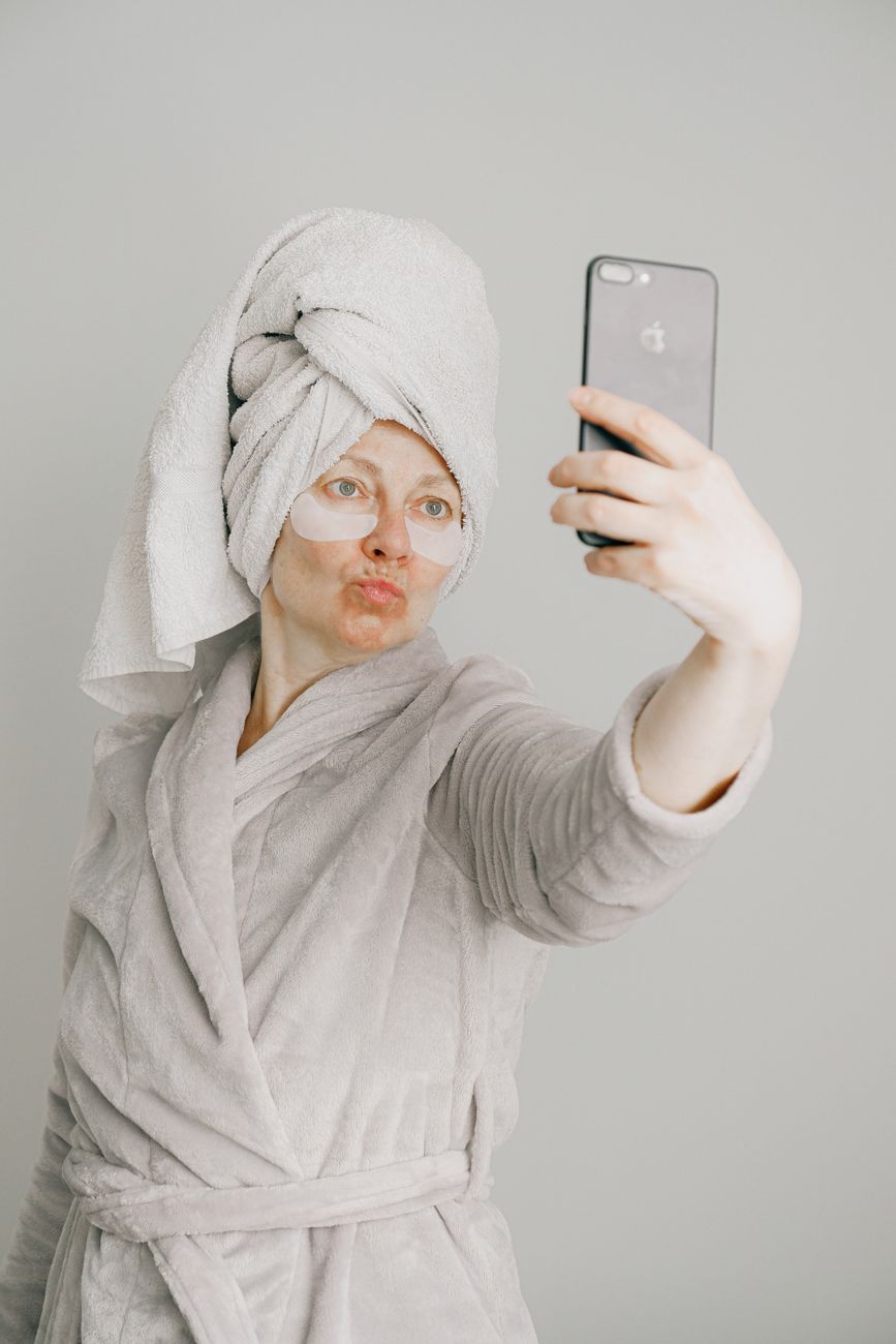 A woman in a bathrobe with an eye mask applies and takes a selfie in a bright, minimalist room.