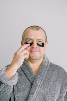Adult caucasian man in a bathrobe applying black under-eye patches for skincare.