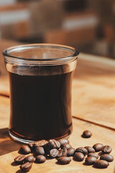 Black coffee served in a clear glass with scattered coffee beans on a wooden table in natural daylight.