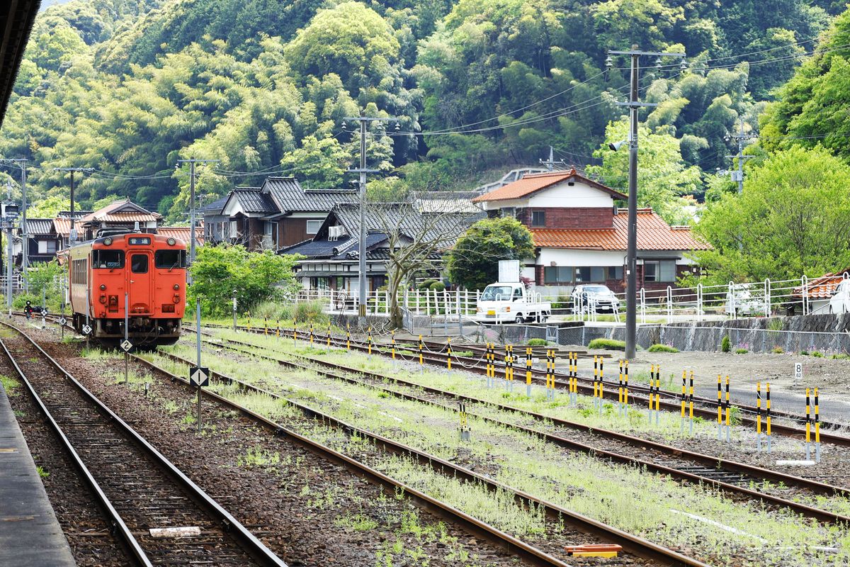 A red local diesel train arriving at Tsuwano Station with green hills behind