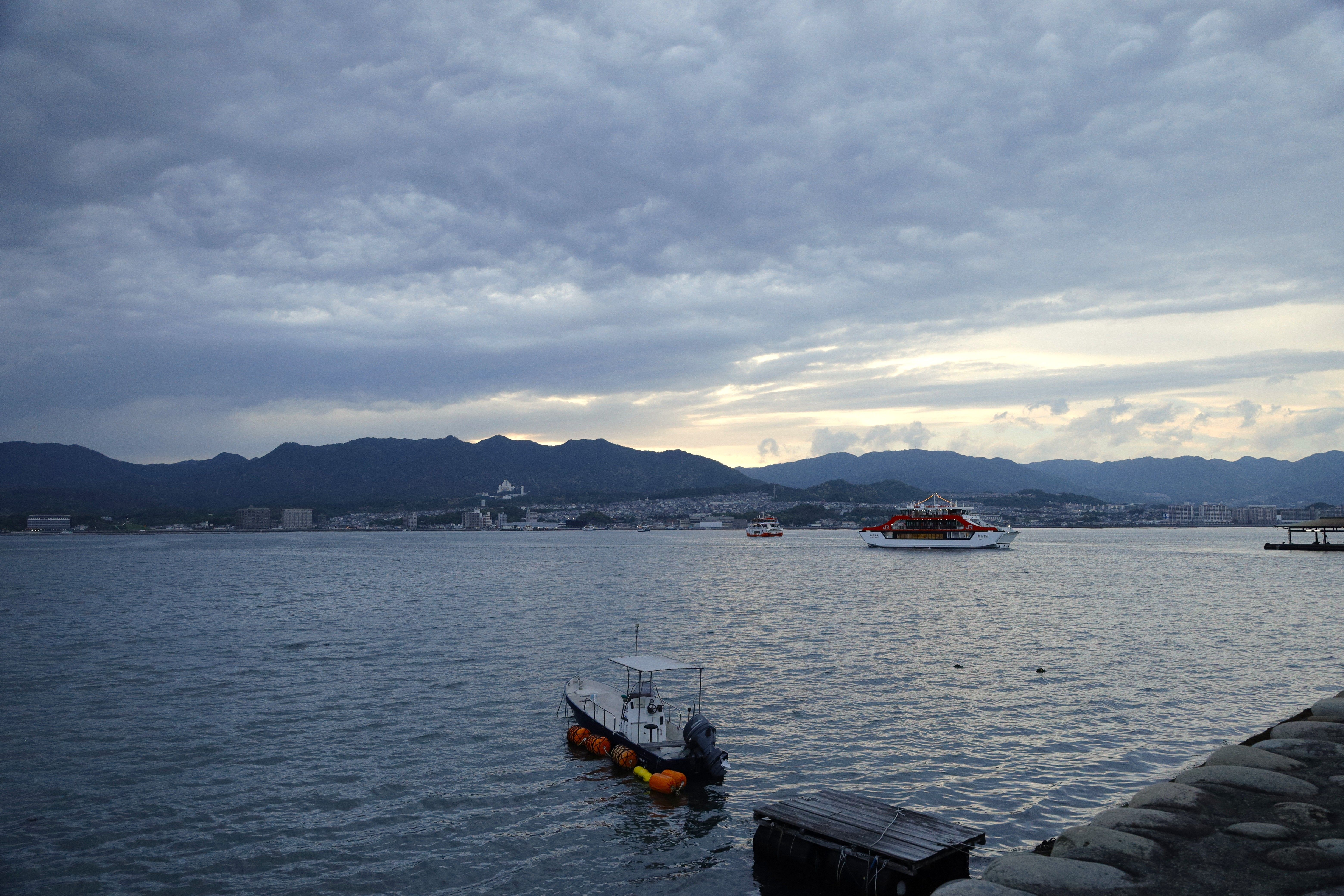 A serene view of Hiroshima's coastline with boats and mountains under a cloudy sky.