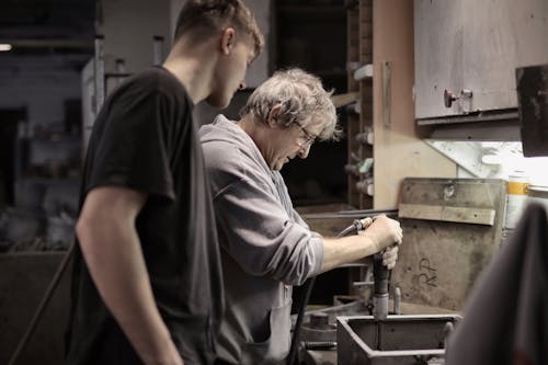 Side view of senior artisan in eyeglasses handling detail in workshop while apprentice looking at process