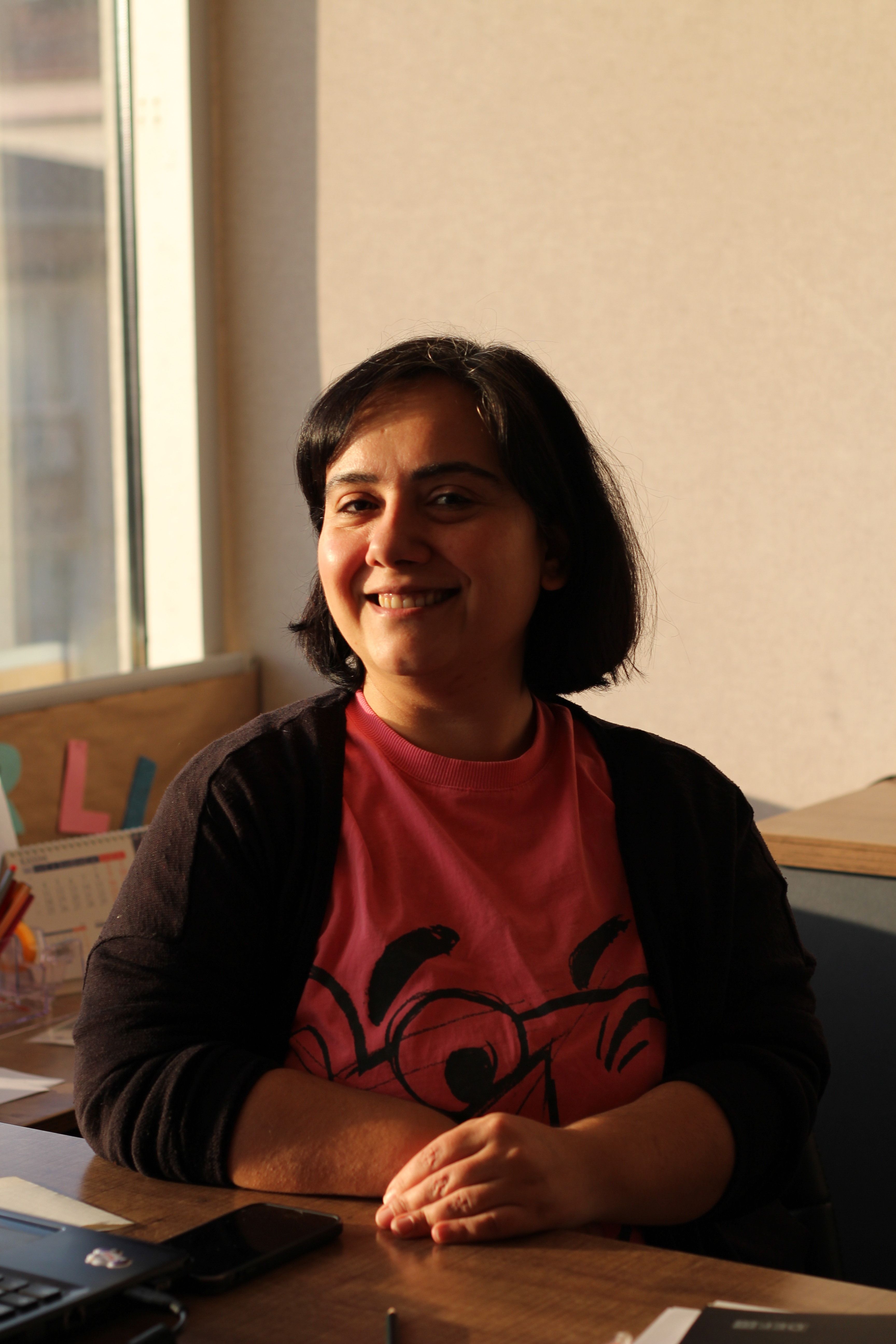 A woman smiling while sitting in a sunlit indoor office environment, wearing a casual shirt.