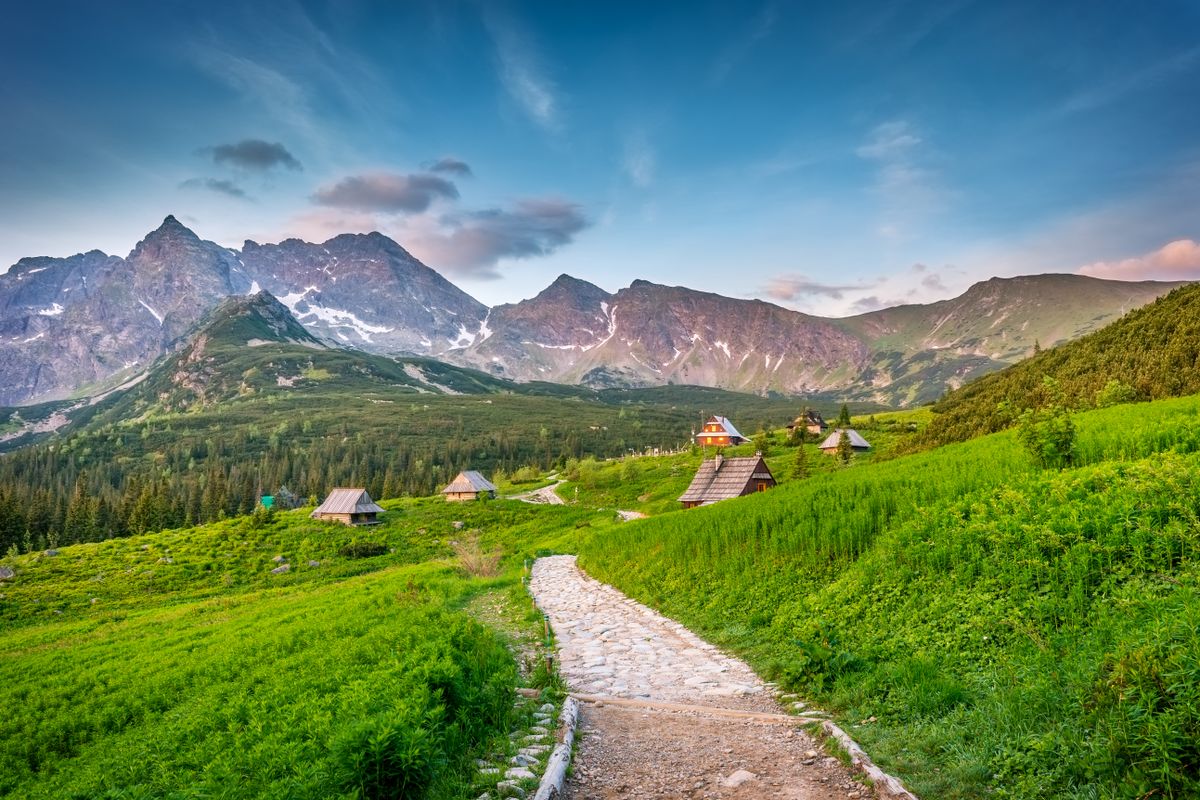 Poteca prin pasune verde cu colibe si creste in fundal