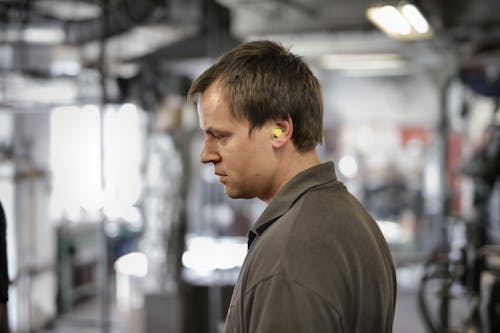Side view of serious young male fitter in earplugs and dark grey tee shirt standing in workshop