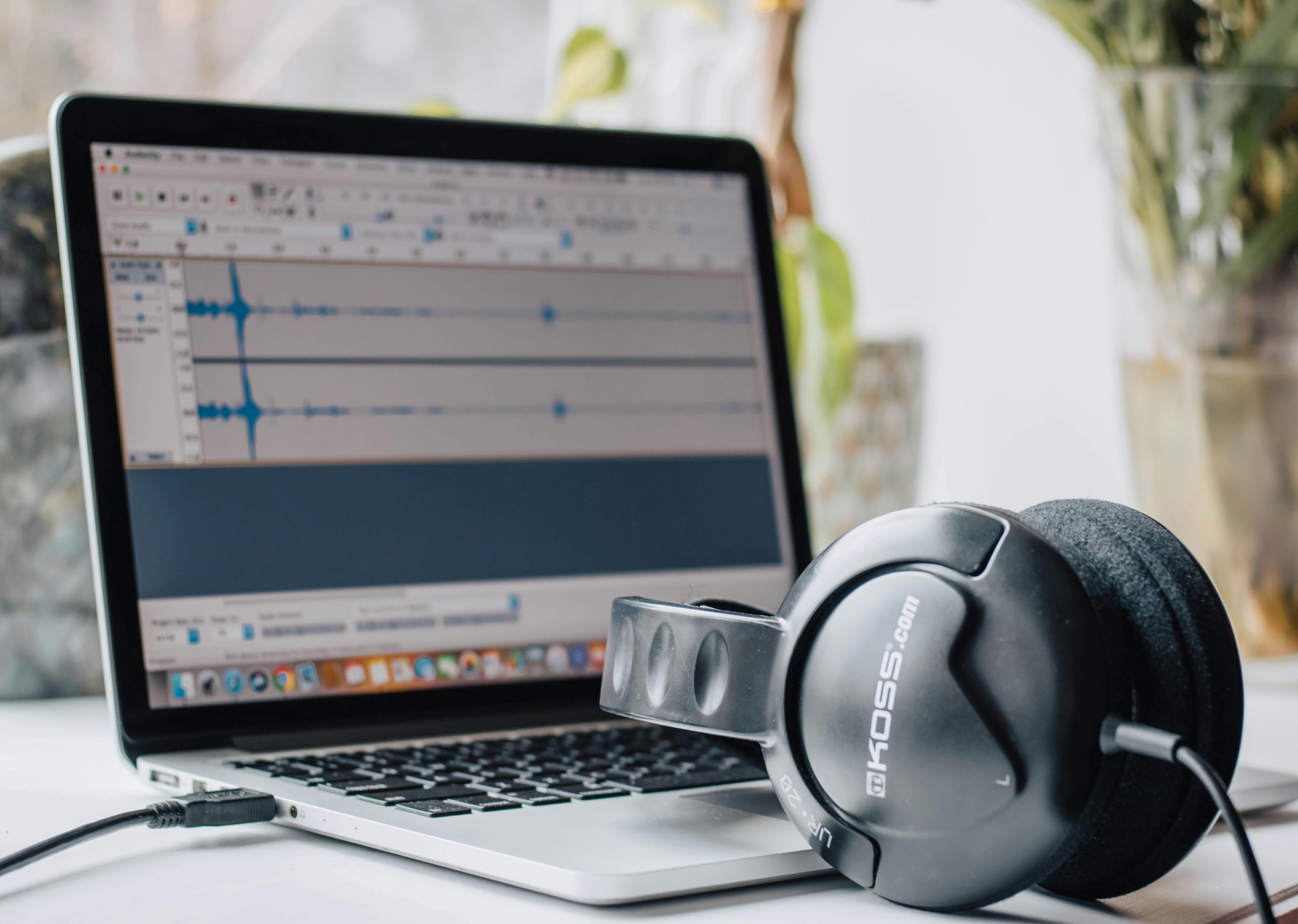 Close-up of a laptop with audio editing software and headphones in a home office setting.