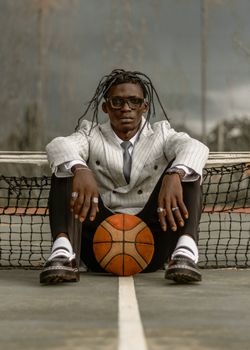 A fashionable man in a suit sits with a basketball on an outdoor court.