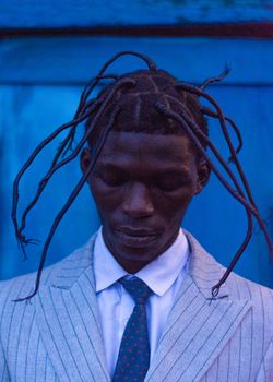 A portrait of a stylish person with unique braided hairstyle against a blue background.