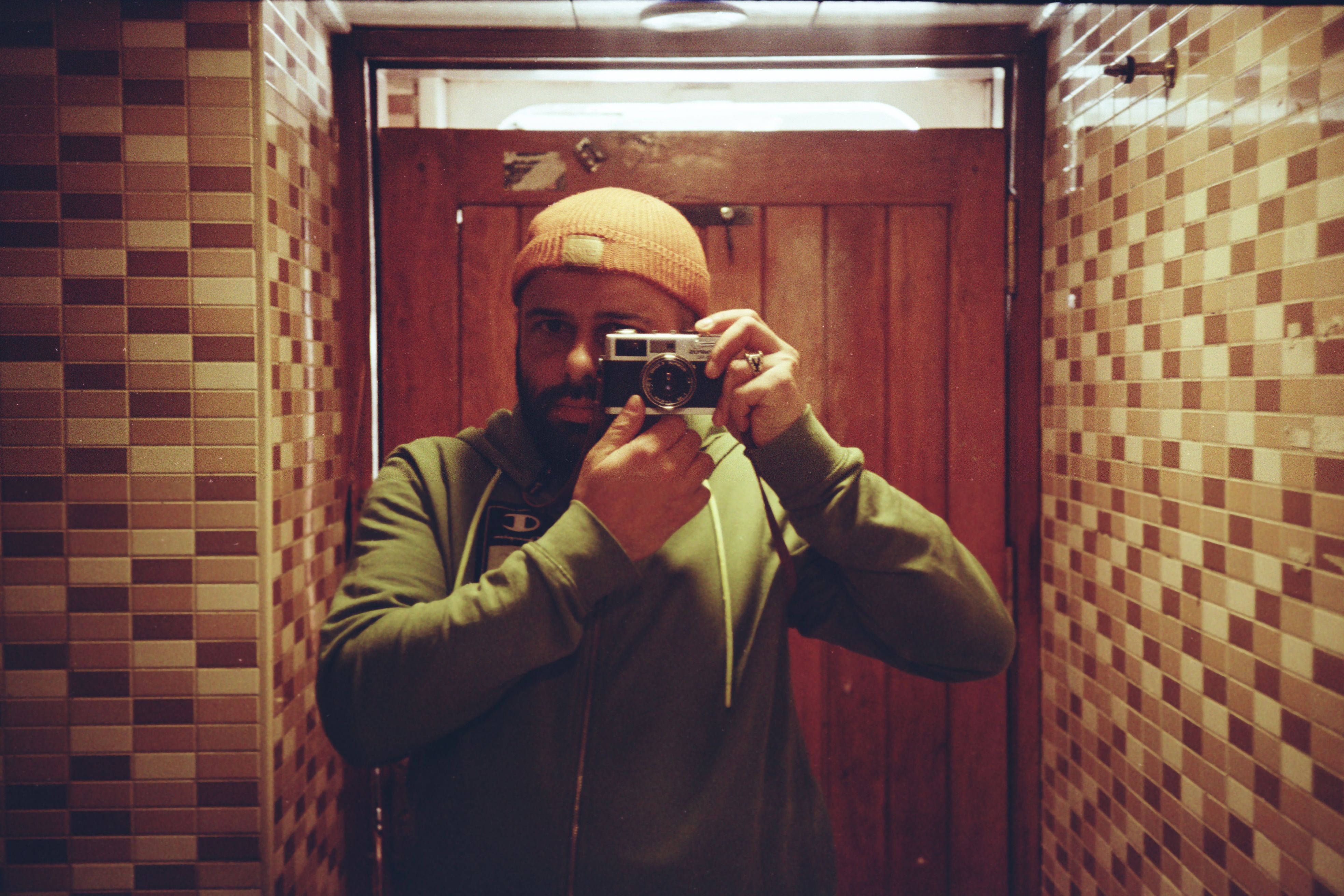 A man wearing a beanie takes a self-portrait with a vintage camera in a tiled room.