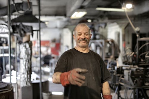 Cheerful senior white hair craftsman in heavy duty gloves laughing in workshop while looking at camera