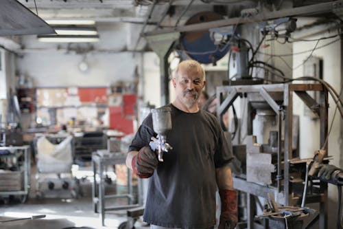 Pleased senior white hair craftsman in heavy duty gloves showing sandblaster and looking at camera