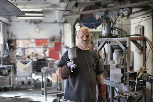 Pleased senior white hair craftsman in heavy duty gloves showing sandblaster and looking at camera