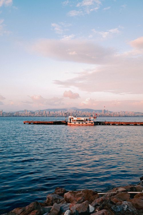 Free Boat docked on a calm sea with city skyline at dusk, under a pastel sky. Stock Photo