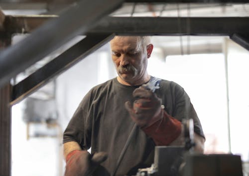 Elderly white hair craftsman with mustache in heavy duty gloves attentively working in workshop