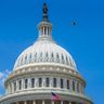 A military fighter jet flies past the dome of the United States Capitol building under a clear blue sky.