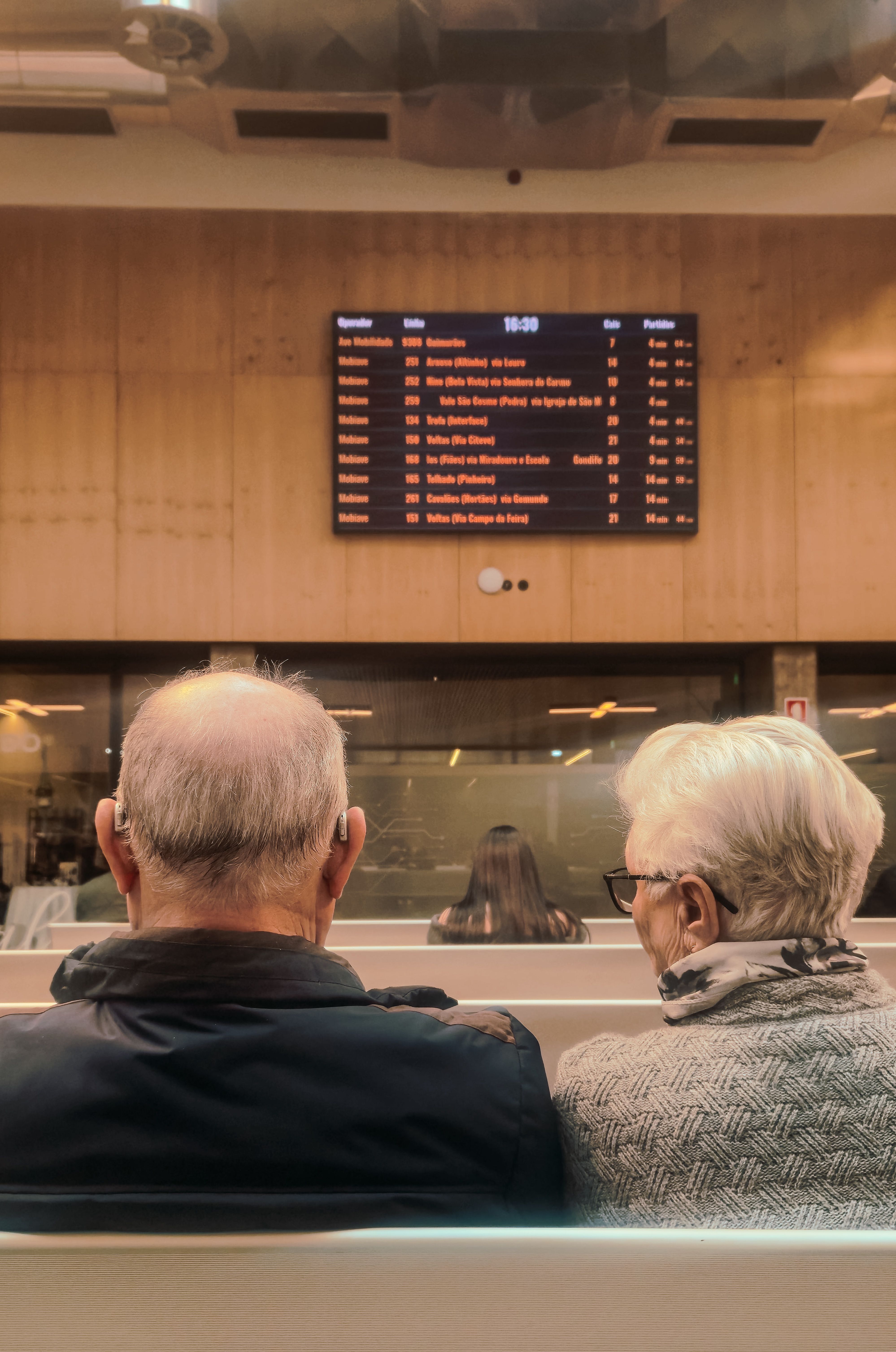 An elderly couple sitting in a train station waiting area with a timetable screen in view.