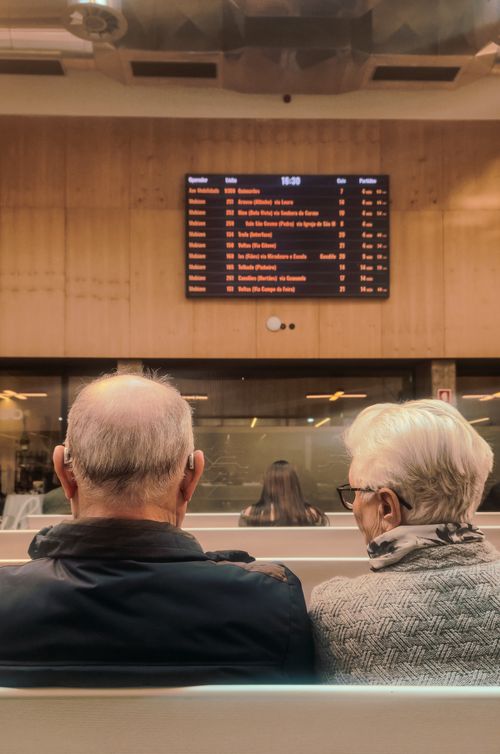 Free An elderly couple sitting in a train station waiting area with a timetable screen in view. Stock Photo
