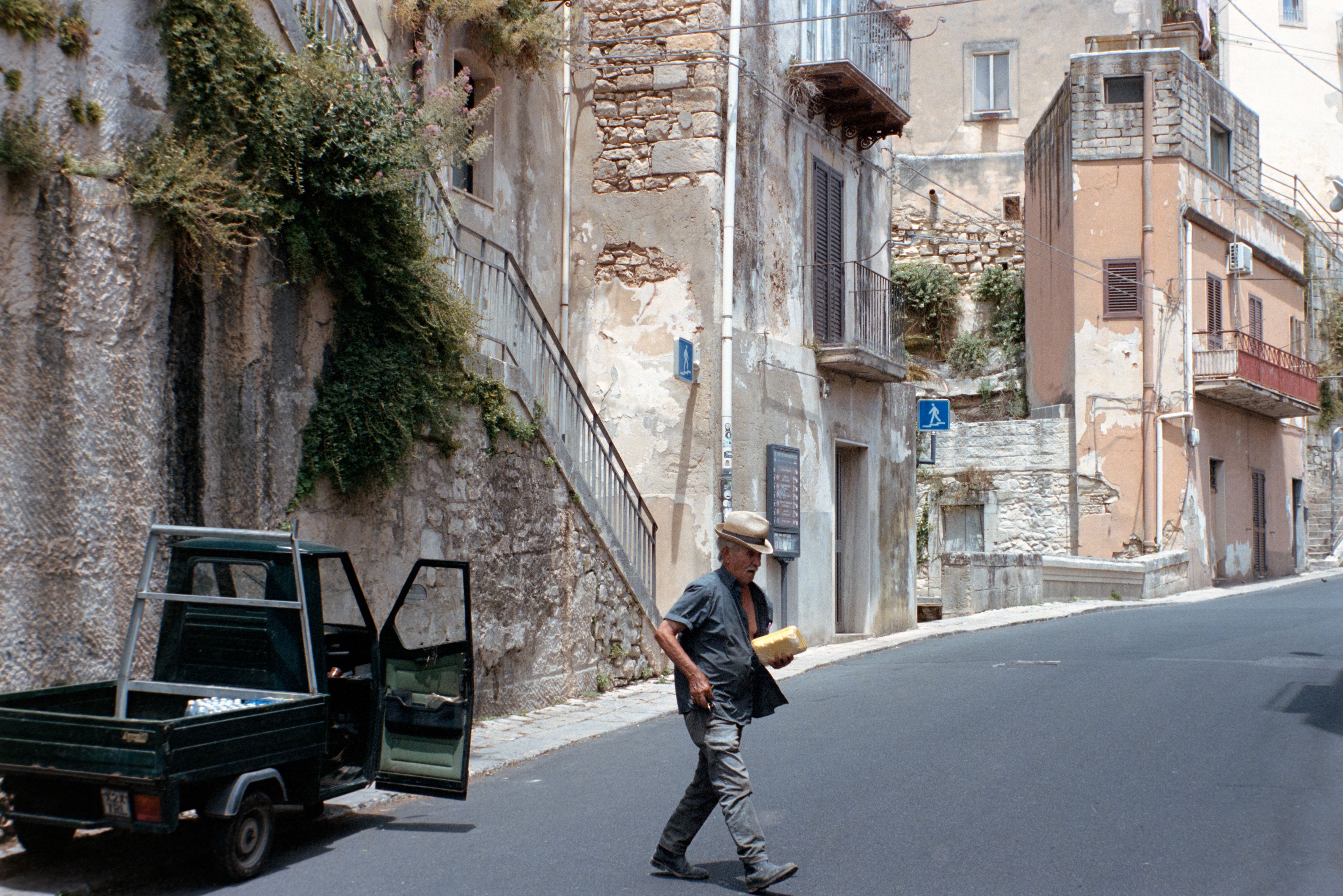 A man crosses a street in a rustic Sicilian village, showcasing ancient architecture and charm.