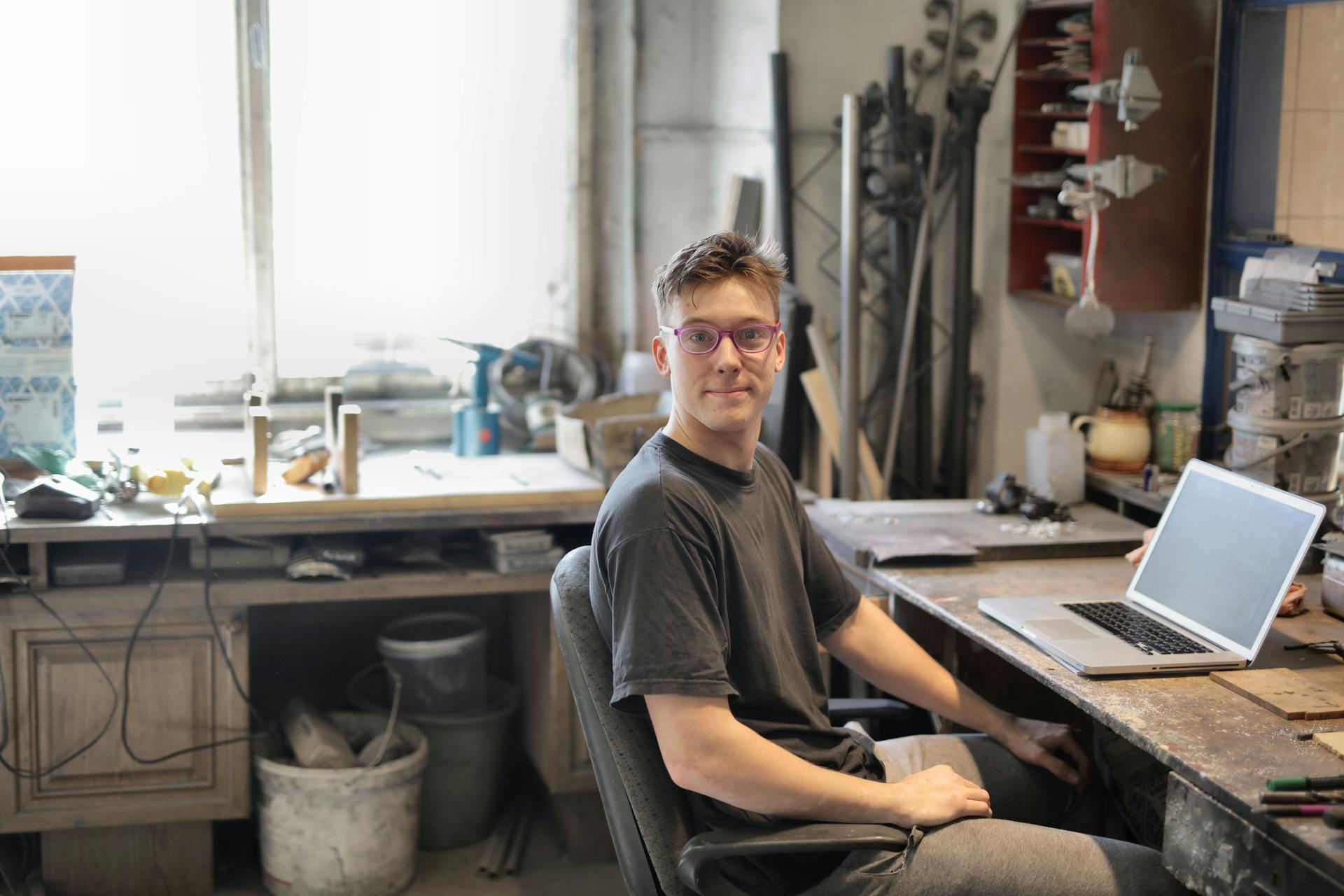 A young man sits in an industrial workshop using a laptop for remote work.