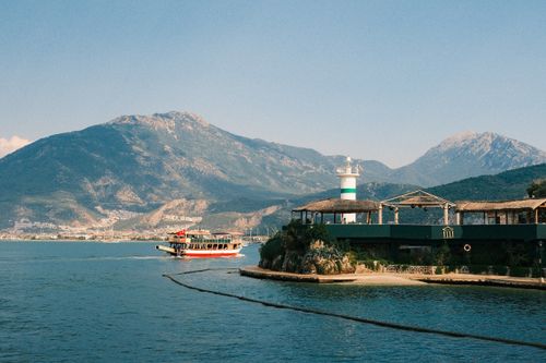 Free A serene landscape featuring a ferry and lighthouse against a mountainous backdrop on a sunny day. Stock Photo
