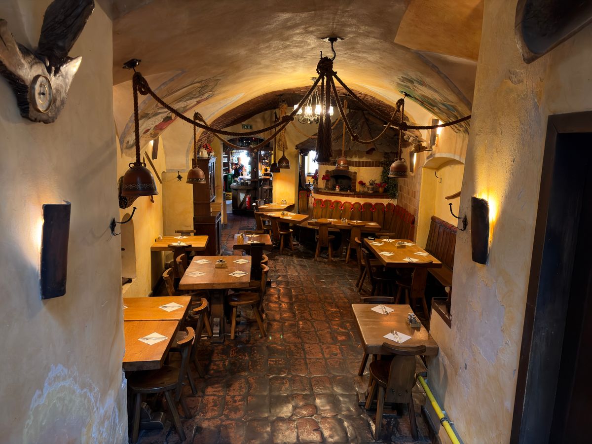 Interior de tavernă în pivniță boltită de piatră, cu mese și bănci simple de lemn și lămpi calde