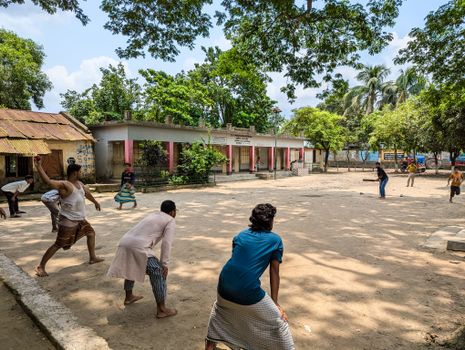 Group of men playing casual cricket in a rural schoolyard on a sunny day.