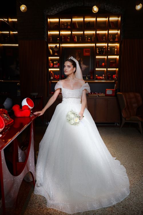 Free A beautiful bride in a white gown holds a bouquet indoors, with stylish decor. Stock Photo